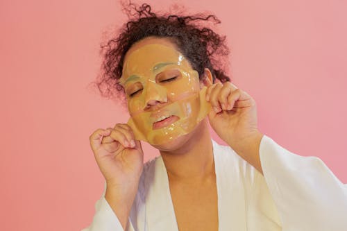 Young black female with curly hair in robe removing hydrogel sheet mask from face with closed eyes