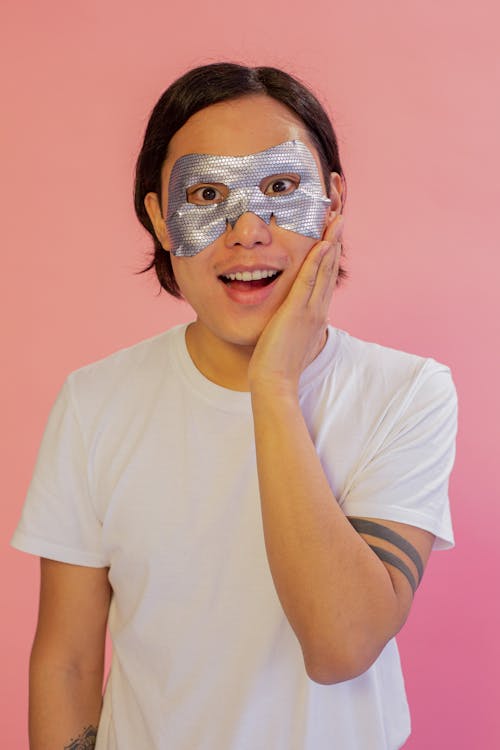 Surprised Asian man with sheet mask touching cheek
