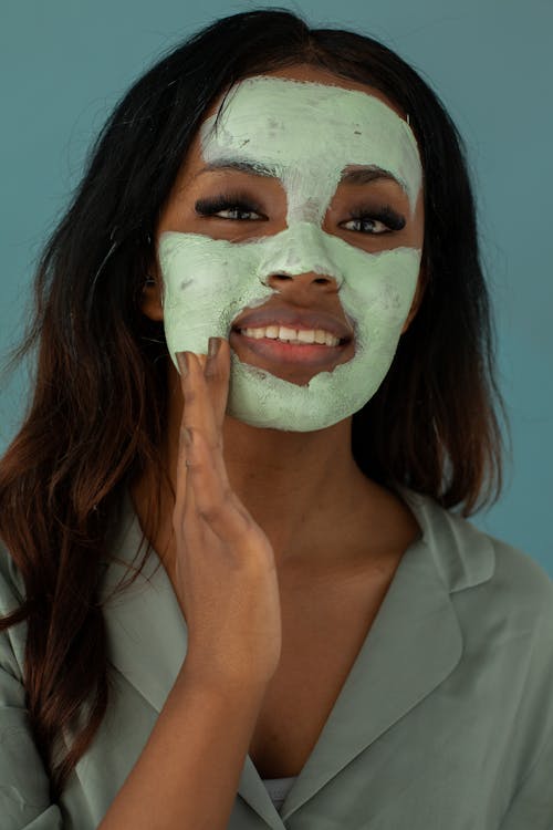 Cheerful young African American female applying mask on face while looking forward on blue background