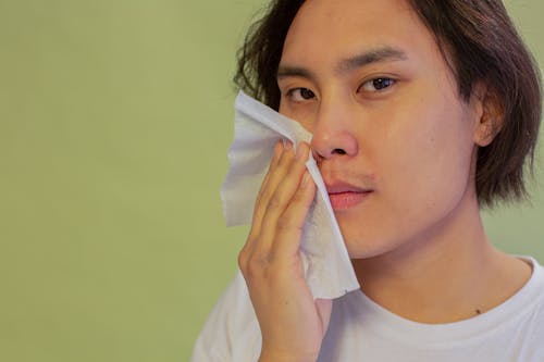 Crop young ethnic male with smooth face skin touching cheek with cleansing wipe while looking at camera