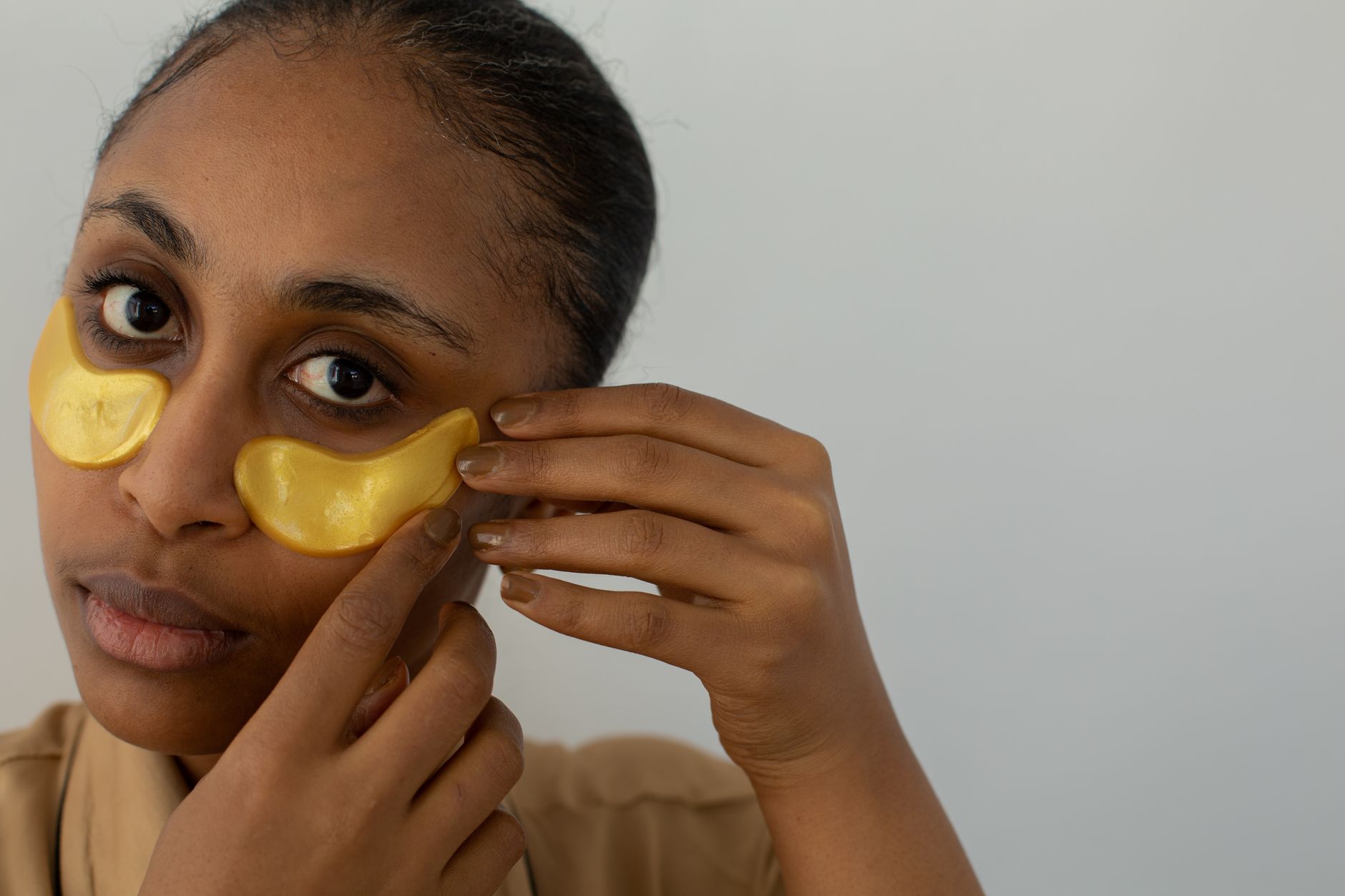 African American female applying hydrogel eye patch on face while looking at camera on light background