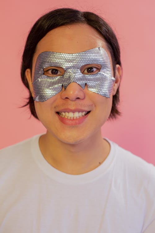 Cheerful ethnic male in t shirt with hydrating eye mask on face looking at camera
