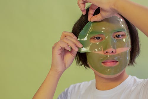 Young unemotional Asian male in white shirt removing cleansing mask sheet from face and looking at camera against green background