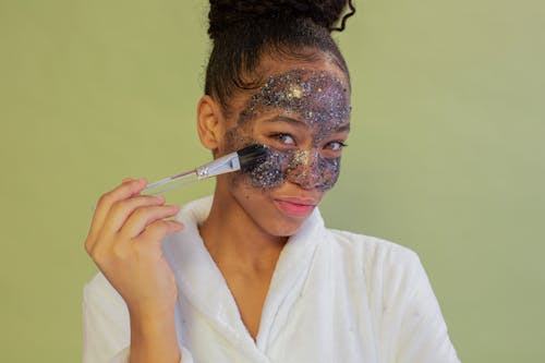 Positive black woman applying sparkling face mask with brush