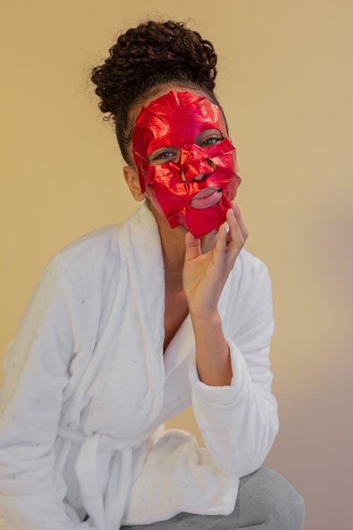 Young glad African American female wearing white bathrobe and red cosmetic face mask sheet touching face and looking at camera contentedly