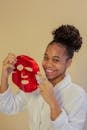 Happy young African American female wearing white bathrobe applying red moisturizing face mask sheet and looking at camera with toothy smile