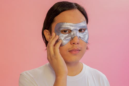 Emotionless young Asian male in white shirt with cosmetic eye mask touching face and looking at camera against pink background
