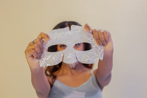 Calm ethnic female showing rejuvenating eye mask sheet in outstretched arms and looking at camera in studio