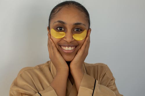 Happy African American female in bathrobe wearing soothing eye patches touching face tenderly and looking at camera with toothy smile against white background