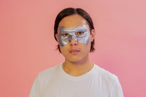 Serious Asian male with moisturizing mask on eyes looking at camera on pink background during skincare procedure in light studio