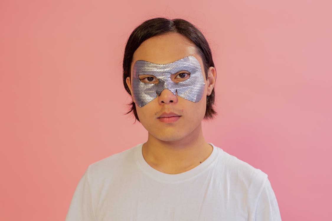 Serious Asian male with moisturizing mask on eyes looking at camera on pink background during skincare procedure in light studio