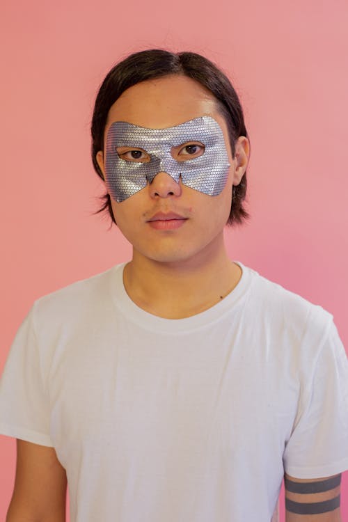Asian male wearing white t shirt and eye mask for skincare looking at camera on pink background in light studio