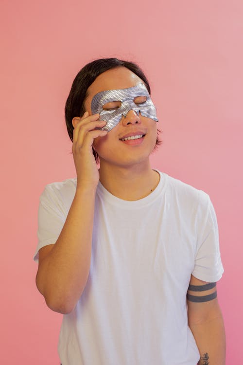 Content Asian male with closed eyes wearing eye mask standing on pink background during beauty skincare procedure in light studio