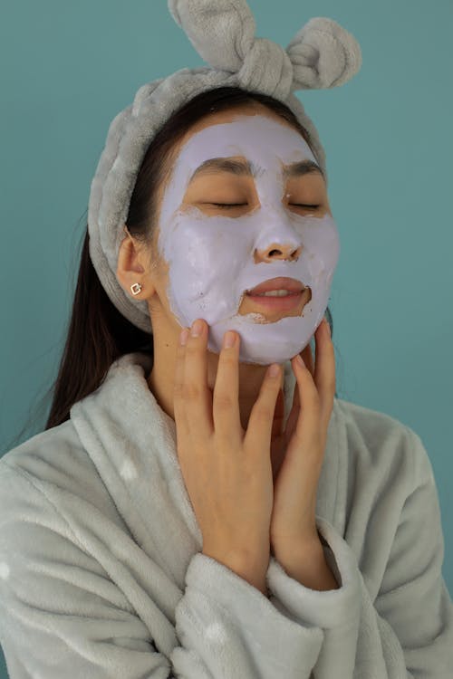 Tranquil female with closed eyes and facial mask wearing bathrobe while standing on blue background during beauty procedure in studio