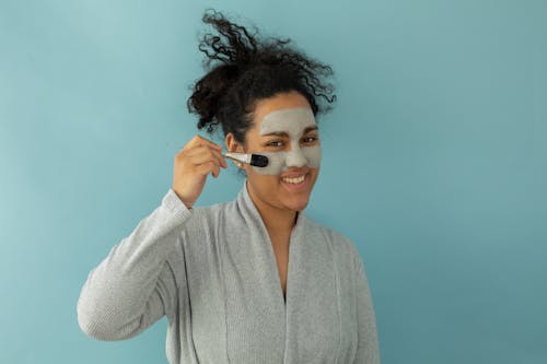 Cheerful African American female in bathrobe applying moisturizing facial mask with brush while looking at camera on blue background during skincare routine