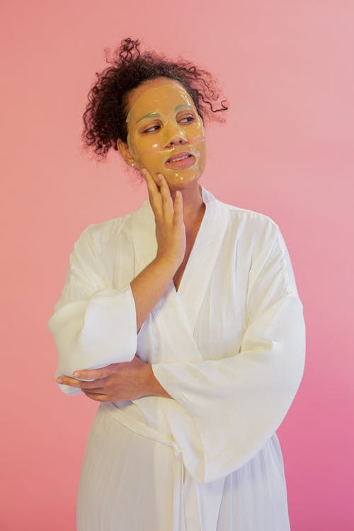 Free Thoughtful African American female in bathrobe with mask on face looking away while standing on pink background during skincare procedure Stock Photo