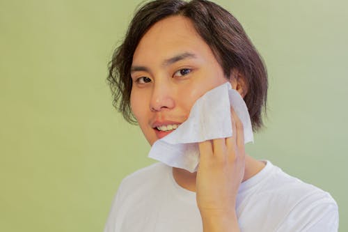 Cheerful Asian male wiping face with wet wipe and looking at camera on green background during hygienic routine in studio