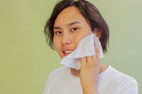 Free Cheerful Asian male wiping face with wet wipe and looking at camera on green background during hygienic routine in studio Stock Photo