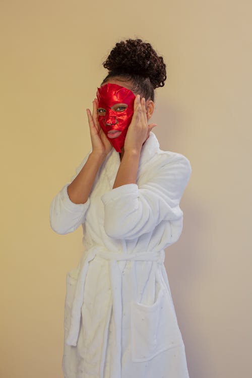 Young African American female with red mask on face wearing white bathrobe and looking at camera on beige background during skincare routine