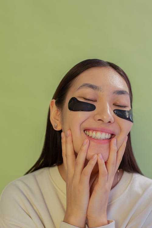 Happy young Asian female with closed eyes and black eye patches touching face while standing on green background in studio