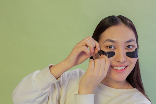 Free Optimistic Asian female wearing white wear applying black moisturizing eye patches while looking away on green background during skincare procedure Stock Photo