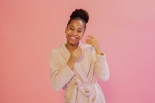 Joyful African American female with lip mask in hands wearing bathrobe while looking at camera on pink background during skincare procedure