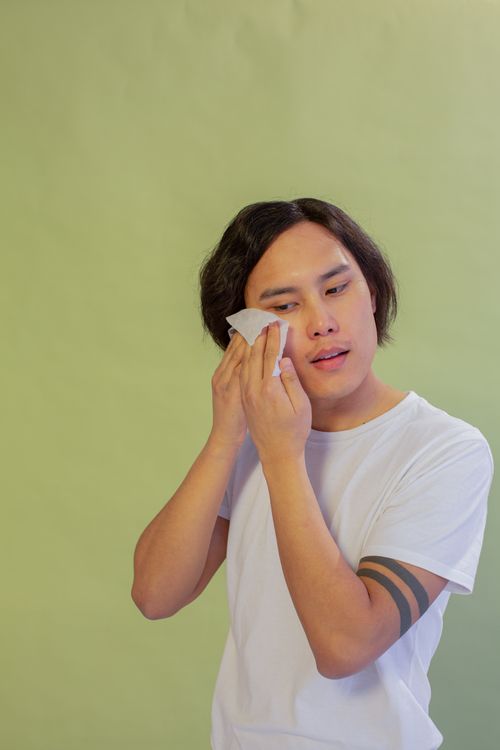 Free Calm Asian male wiping face with wet wipe while standing on green background during daily hygienic procedure in light studio Stock Photo