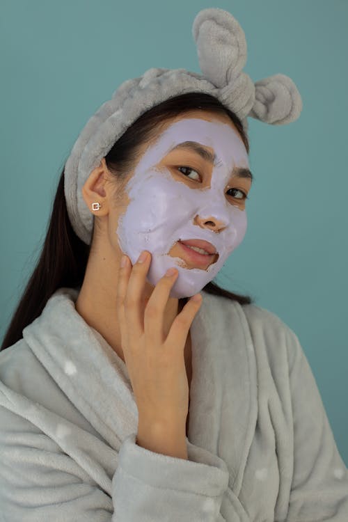 Content Asian female wearing bathrobe with facial mask for daily skincare treatment looking at camera on blue background in studio