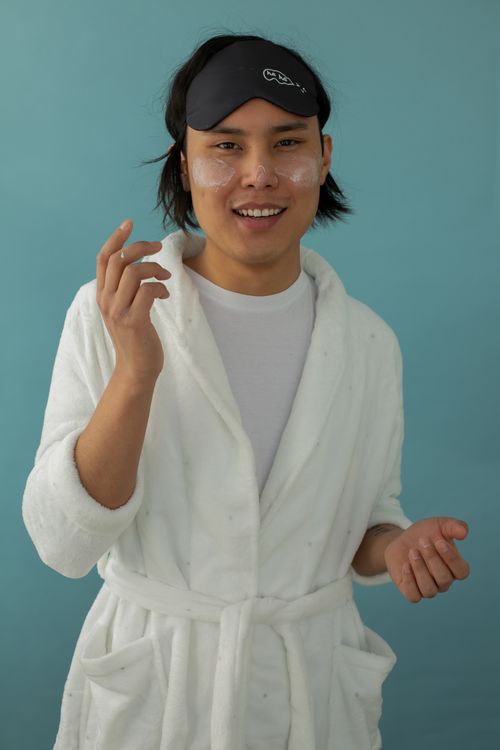Free Portrait of a young man in a white bathrobe wearing a sleep mask and under eye patches against a blue background. Stock Photo