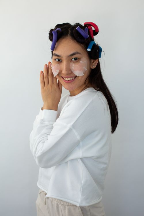 Free Portrait of an Asian woman wearing hair rollers and eye patches, posing with a smile in a white background. Stock Photo