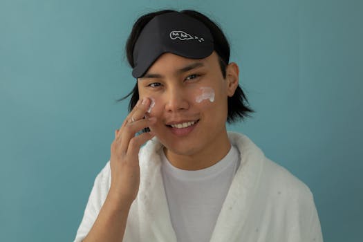 Smiling man in robe applying facial cream with sleep mask on a blue background.