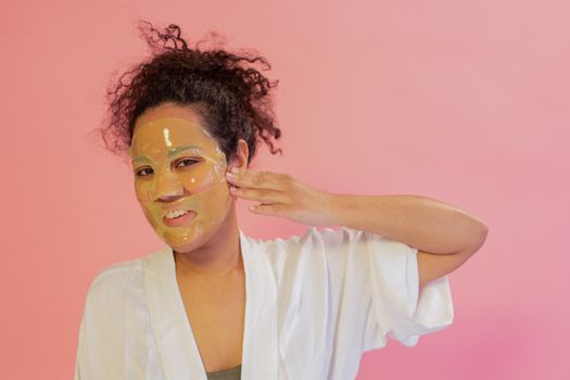 African American woman applying face mask on pink background, emphasizing skincare routine.
