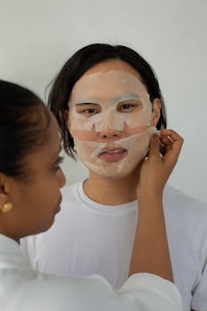 Woman applying a sheet mask to another woman's face in a skincare routine setting.