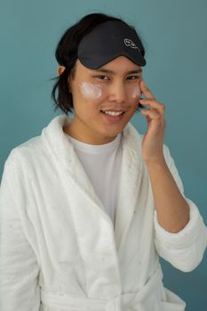 Asian man in robe applying skincare cream with a sleep mask in studio setting.