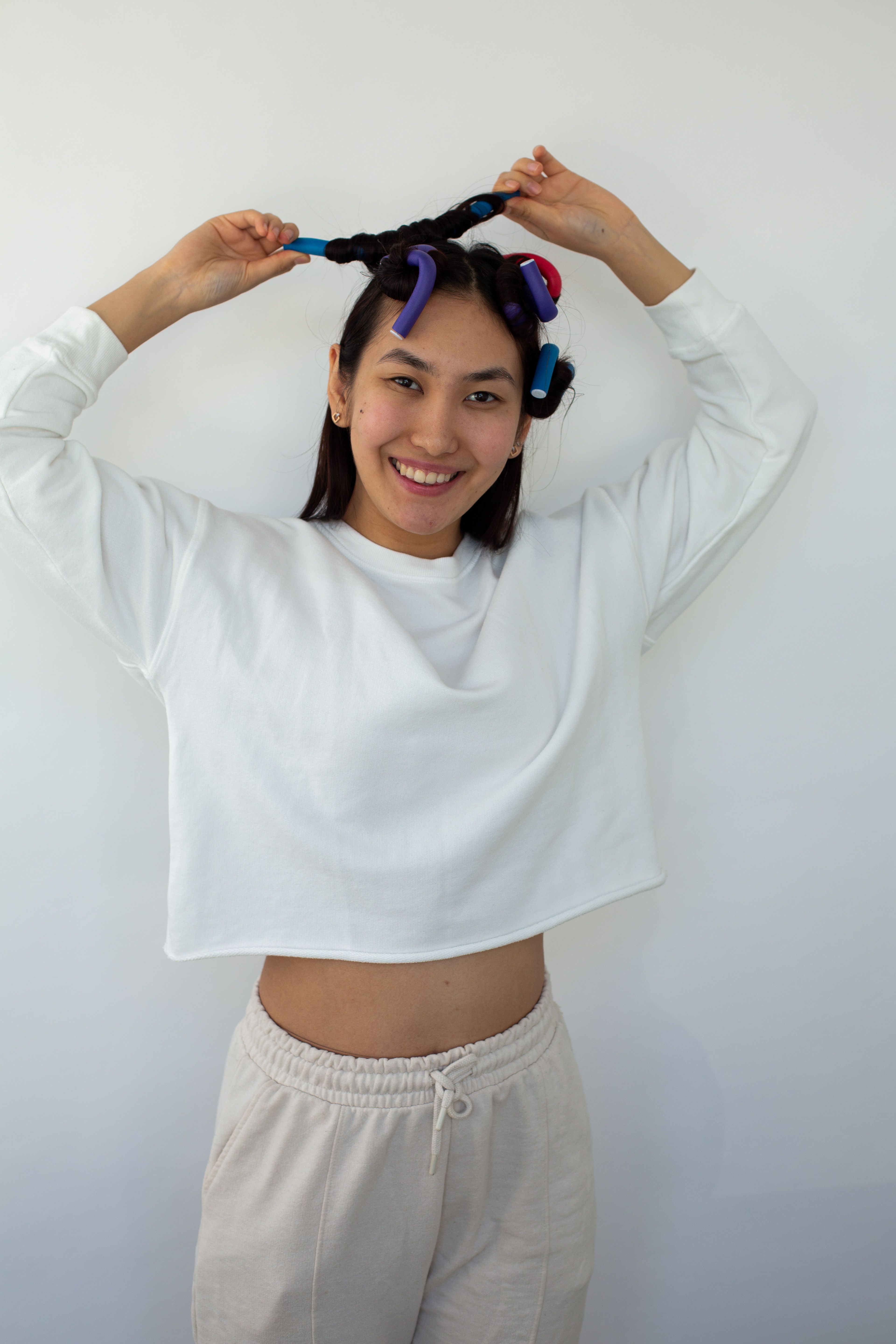 Joyful young woman in a white jumper putting rollers into her hair against a white wall