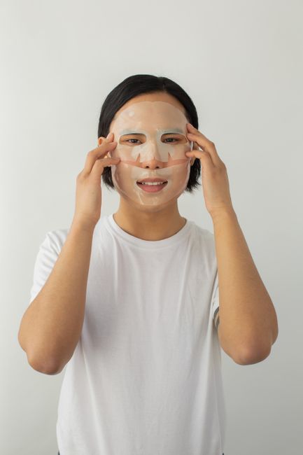 Young ethnic male touching face while applying sheet mask and looking at camera on light background