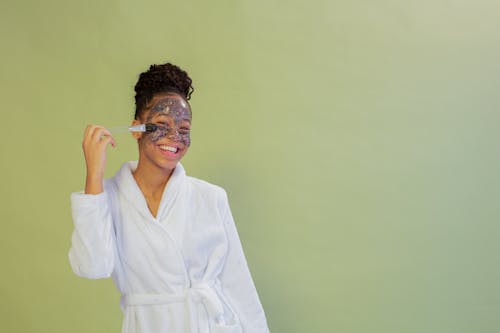 Content African American female teenager in bathrobe applying exfoliating mask on face while looking at camera on green background