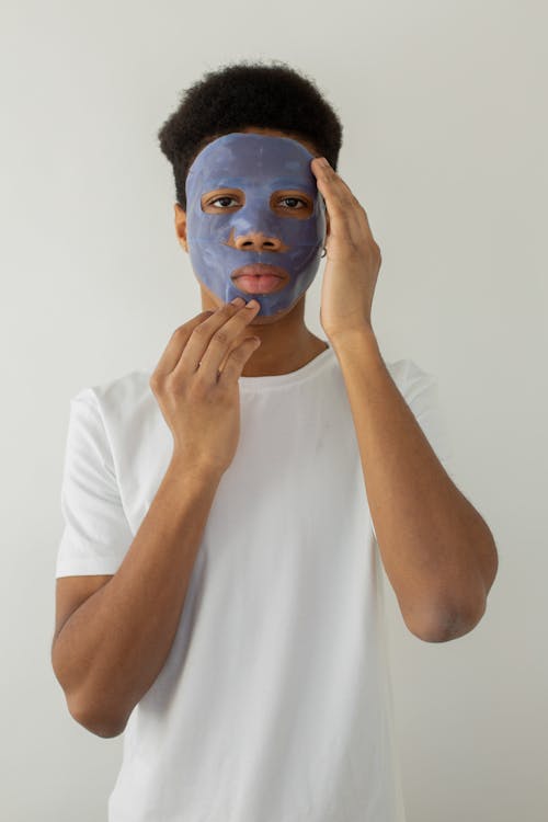 Young black male in t shirt touching chin while applying hydrating mask and looking at camera on light background