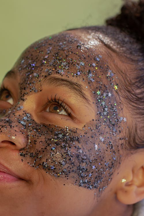 Free Crop black female adolescent looking up with exfoliating mask on cheeks and forehead with stars and shiny particles on green background Stock Photo