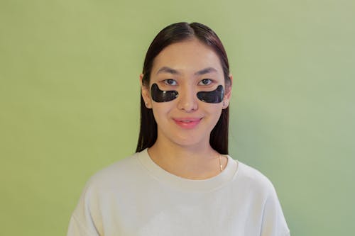 Cheerful ethnic female teenager with hydrogel eye patches on cheeks looking at camera on green background