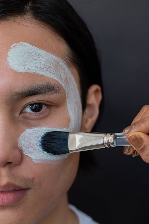 Crop unrecognizable female beautician with brush applying purifying mask on face of ethnic man looking at camera on gray background
