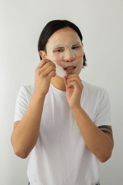 Young ethnic male in t shirt removing hydrating mask from face while looking at camera on light background