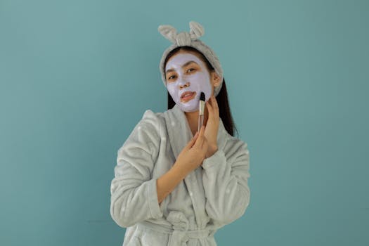 Young ethnic female in robe and headband applying cleansing mask with cosmetic brush while looking at camera on turquoise background