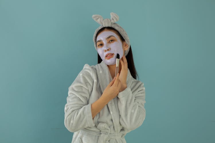 Tender Asian Woman Applying Clay Mask On Face With Applicator