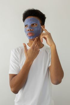 Contemplative ethnic male in t shirt touching face while applying hydrating mask and looking away on light background