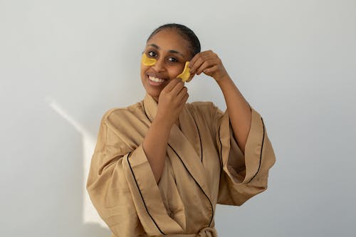 Cheerful African American female in robe applying hydrating eye patches on cheeks while looking at camera on light background