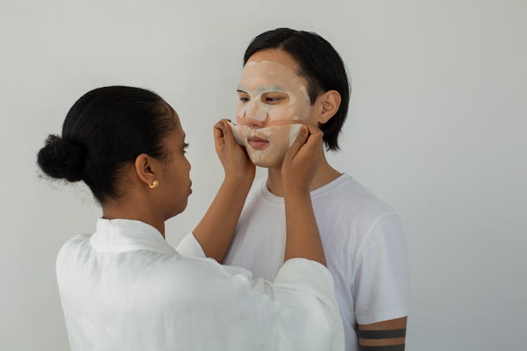 Ethnic Beautician Applying Sheet Mask On Face Of Asian Client