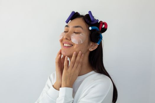 Smiling young ethnic female with hair rollers touching cheeks with eye patches on white background