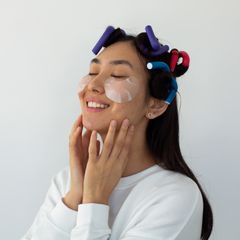 Smiling young woman with dark rollers in her hair and eye patches, hands on her cheeks, on white