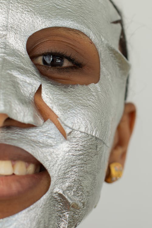 Crop unrecognizable cheerful African American female with gray facial mask looking at camera on white background during daily skincare procedure